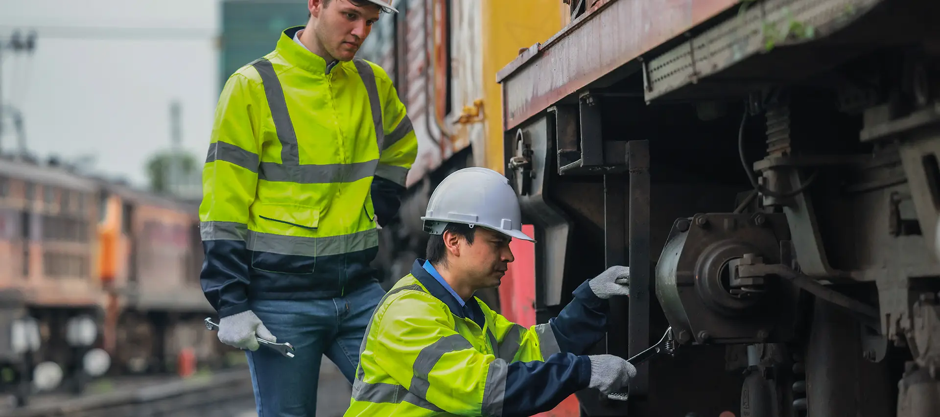Dos operarios realizan tareas de mantenimiento en un vagón. Uno de ellos utiliza una herramienta manual para apretar un perno, mientras que el otro observa el proceso. La imagen pone de manifiesto la necesidad de soluciones de mantenimiento eficientes, fiables y seguras en el sector ferroviario.